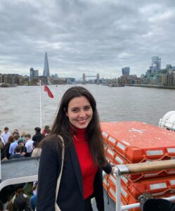 多里·科瓦奇微笑地站在泰晤士河上的一艘船上。伦敦塔桥就在她身后的远处。